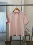 Signature Tee in blush pink on a wooden hanger, showing the blanket-stitch collar and embroidered say logo