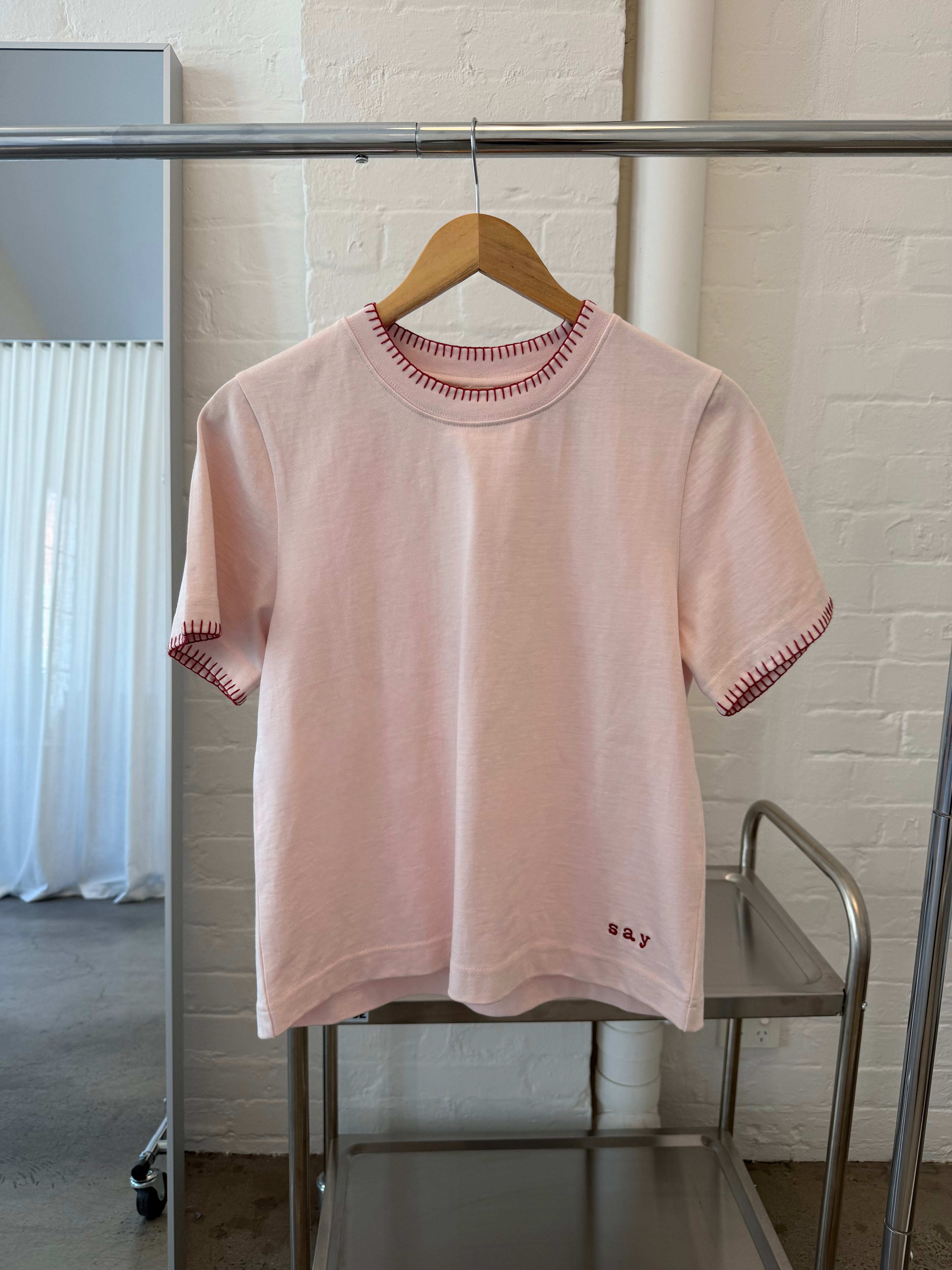 Signature Tee in blush pink on a wooden hanger, showing the blanket-stitch collar and embroidered say logo
